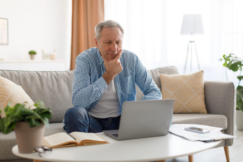 Wenn die Rente nicht reicht Boomer – nachdenklicher Mann 60+ mit Laptop am Schreibtisch, warmes Licht