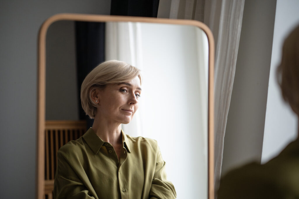 Self-Directed Ageism Boomer – nachdenkliche Frau 60+ vor einem Spiegel, der eine jüngere Version zeigt
