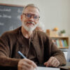 Weiterbildung ab 60 Boomer – selbstbewusster Mann 60+ mit Brille macht sich Notizen in einem Seminarraum mit Tafel und anderen Teilnehmern