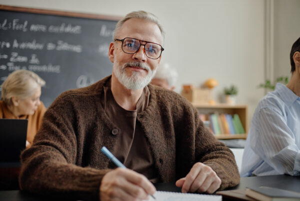 Weiterbildung ab 60 Boomer – selbstbewusster Mann 60+ mit Brille macht sich Notizen in einem Seminarraum mit Tafel und anderen Teilnehmern